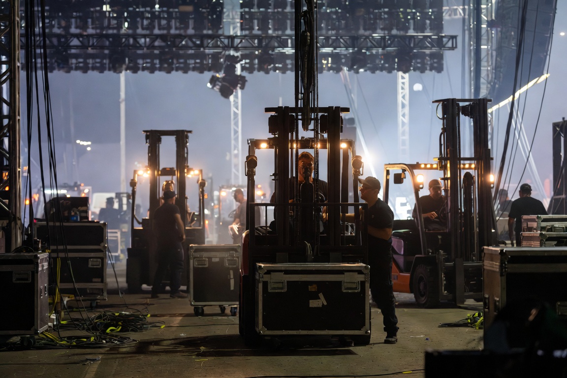 Late-night stage floor thick with haze — several forklifts and crew reduced to silhouettes under backlight.