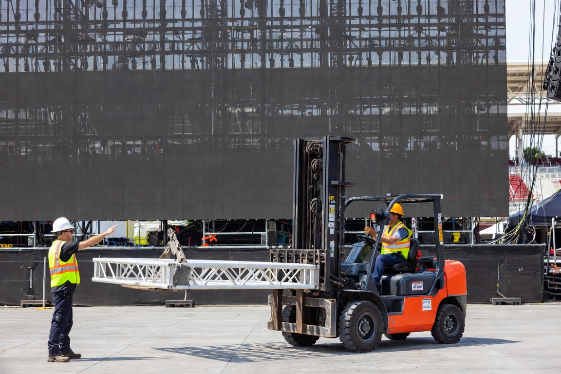 A forklift carries a single truss section while a spotter directs from the ground, LED wall filling the background.