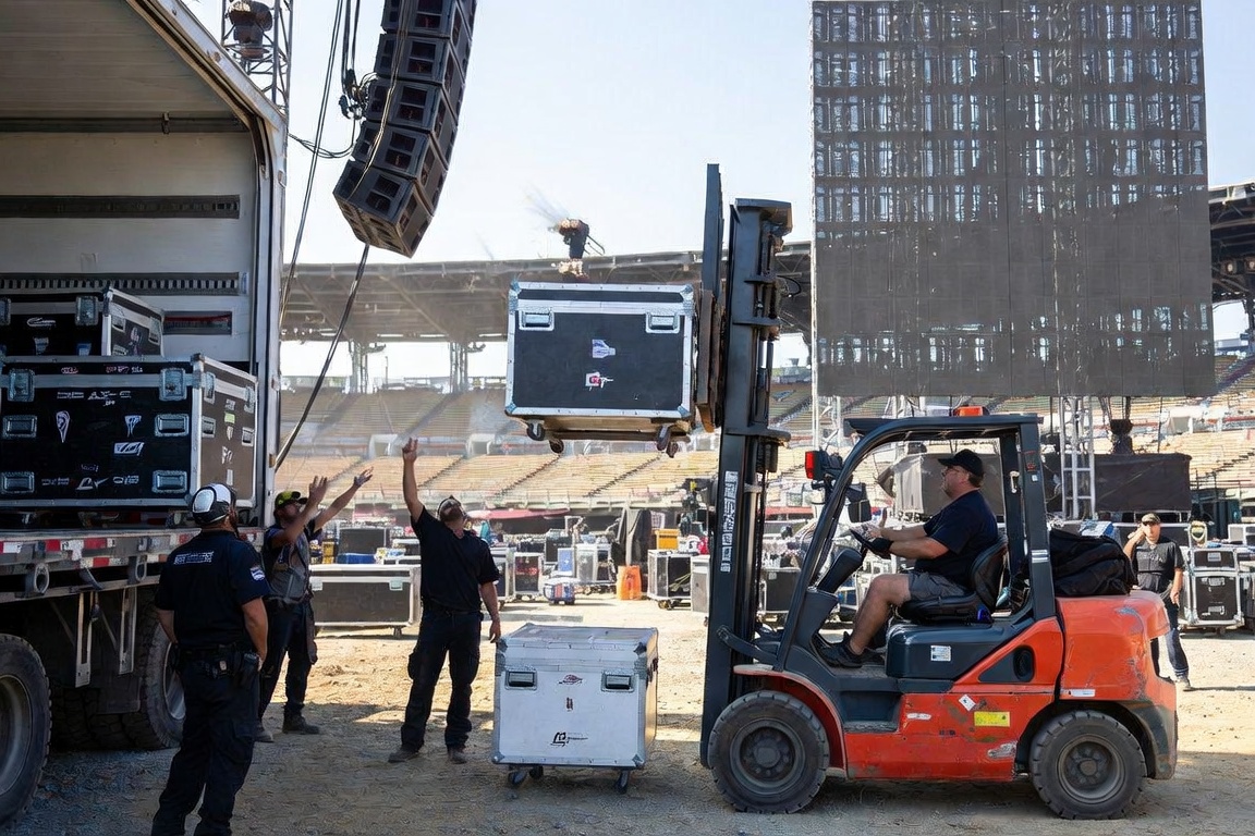 Ground crew with both arms raised, signaling to a forklift operator lifting a road case beside a truck at a daylight stadium build.