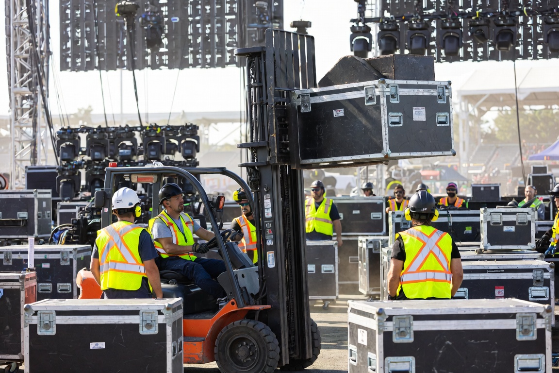 A forklift carries a road case above head height through a dense staging line of cases and hi-vis crew.