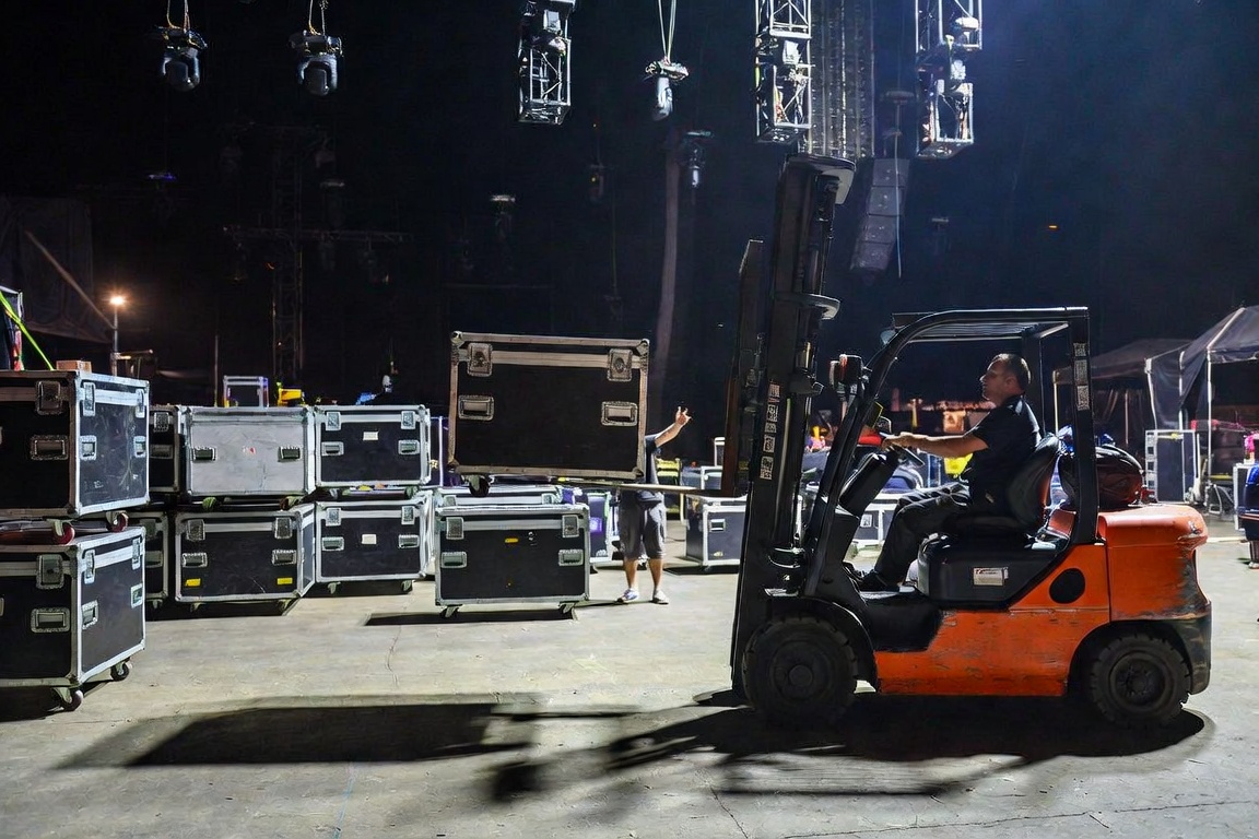 A crew member stands clear of a stack of road cases with one hand raised, signaling to a forklift operator on a darkened stage floor.