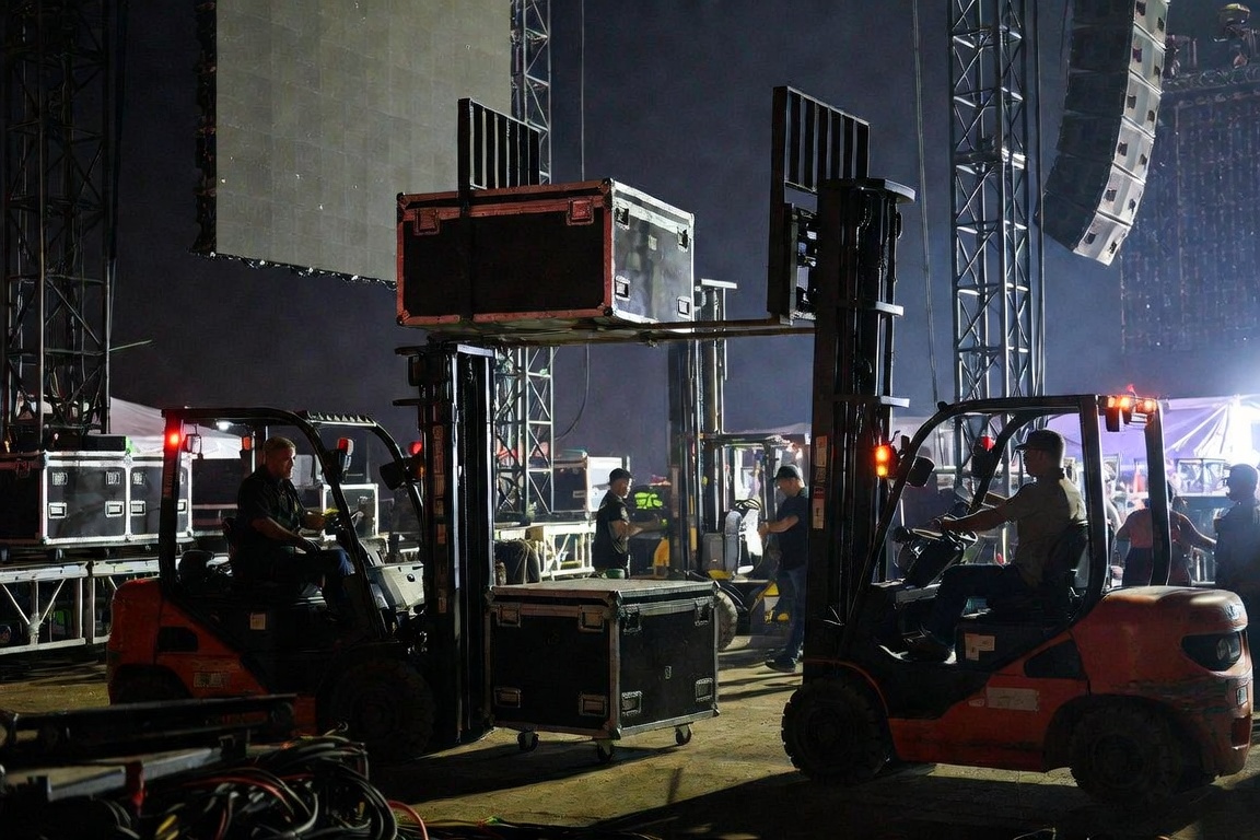 Forklifts carrying road cases at height on a night festival floor, an LED wall and line array behind them.